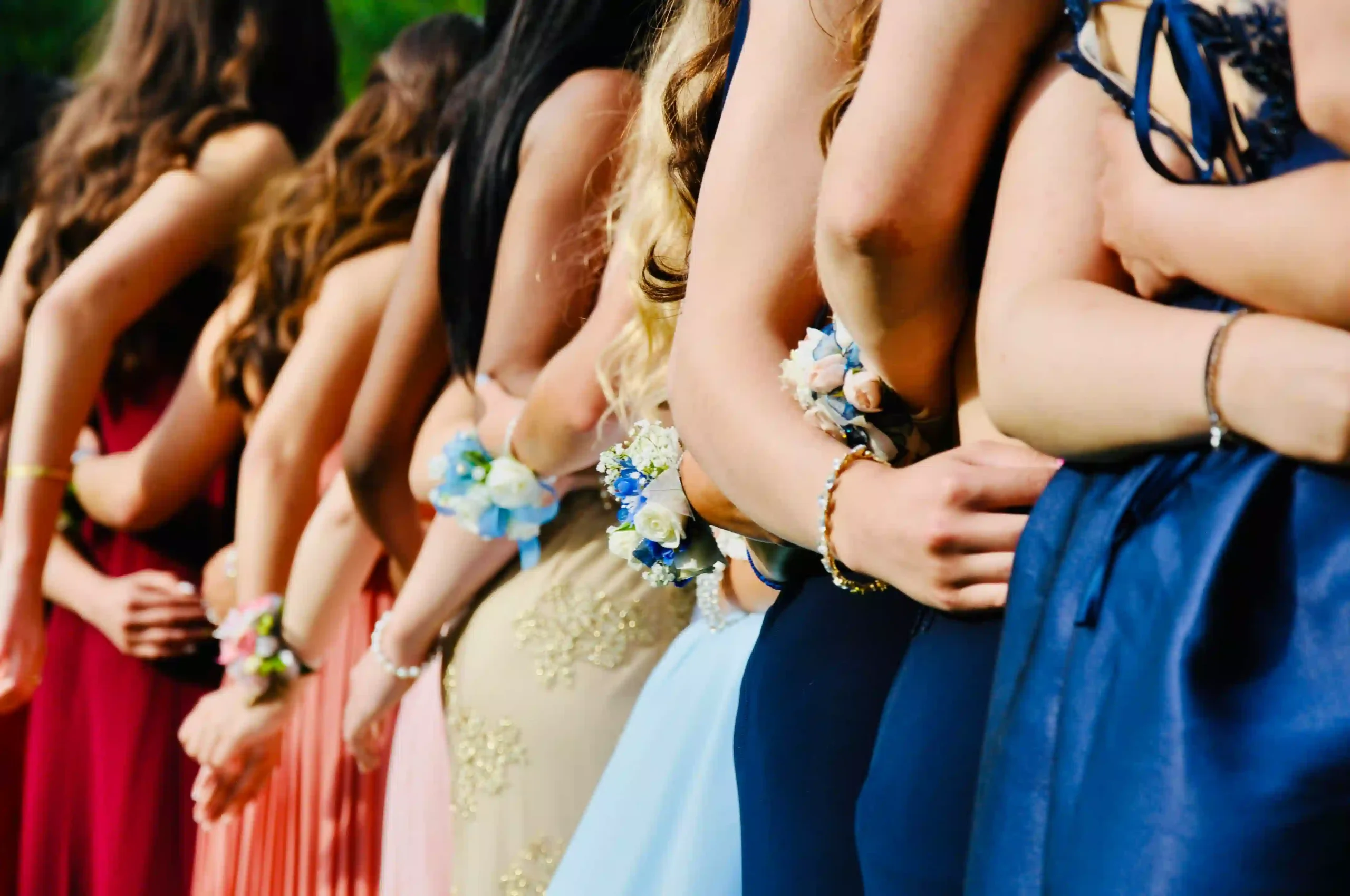 A group of women in formal dresses stand in a line with their arms linked, each adorned with a wrist corsage made of blue and white flowers. The vibrant dresses—dark red, pink, gold, light blue, and navy—evoke images of elegance akin to arriving in style with Bristol Chauffeur Service.