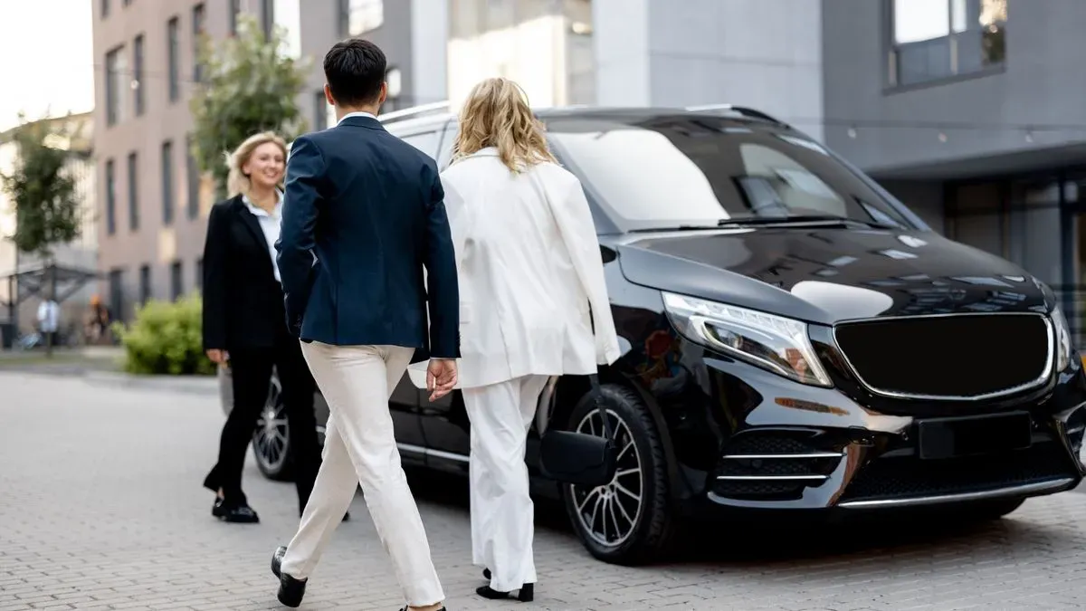 Two people in business attire walk toward a black luxury van parked on a city street, a hallmark of the Bristol Chauffeur Service. A third person in a suit approaches them from the opposite direction, framed by buildings and trees.