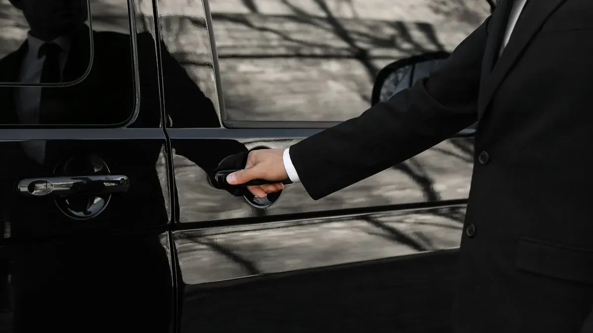 A person in a black suit, reminiscent of a Bristol Chauffeur Service professional, opens the door of a sleek black car. The reflection of their hand glistens on the glossy surface, set against the backdrop of blurred outdoor scenery and tree branches.
